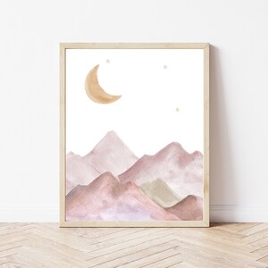 May include: Watercolor print of a mountain landscape with a crescent moon and stars. The artwork features soft, muted tones of pink, brown, and green, set against a white background. The print is framed in a light-colored wooden frame.