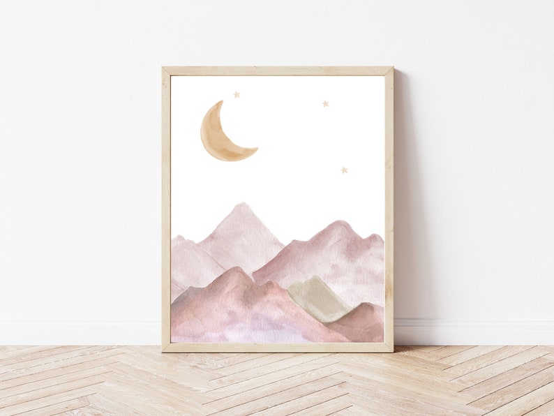 May include: Watercolor print of a mountain landscape with a crescent moon and stars. The artwork features soft, muted tones of pink, brown, and green, set against a white background. The print is framed in a light-colored wooden frame.