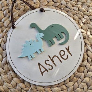 May include: A circular wooden sign with a light wood finish, featuring two dinosaur cutouts in blue and green. The name "Asher" is engraved in a brown font. A brown leather cord is attached for hanging.