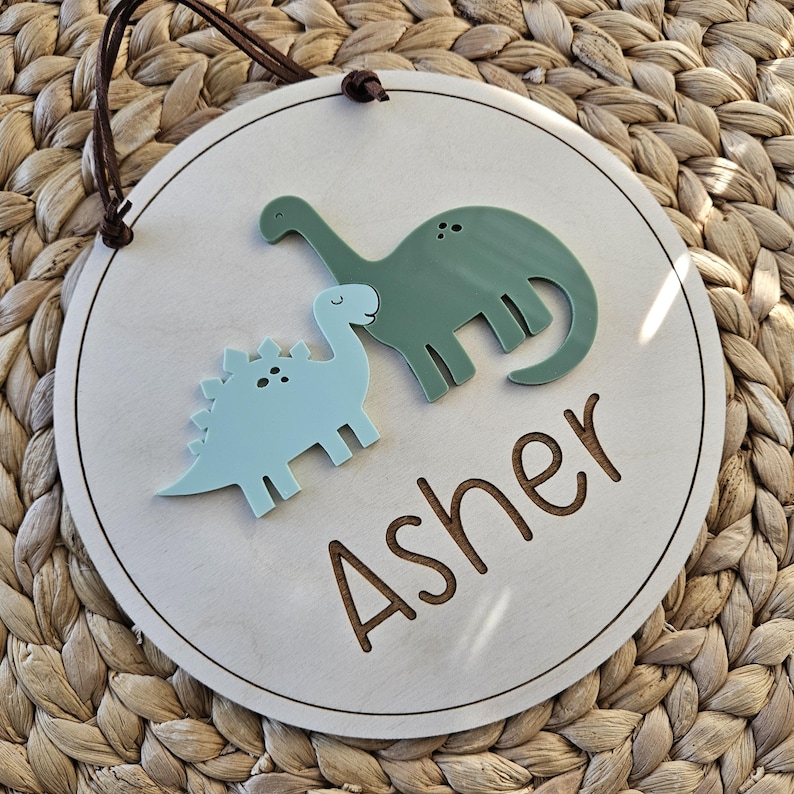 May include: A circular wooden sign with a light wood finish, featuring two dinosaur cutouts in blue and green. The name "Asher" is engraved in a brown font. A brown leather cord is attached for hanging.