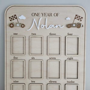 以下が含まれることがあります： 「ONE YEAR OF Nolan」の文字が入った木製のマイルストーンボード。ボードには12個の写真フレーム、レーシングカー、チェッカーフラッグ、1から12までの数字があります。ボードは明るい色の木材で作られています。