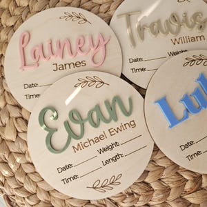 May include: Four circular wooden birth announcement plaques featuring raised acrylic names in pink, sage green, and blue. Each plaque includes laser-engraved text for Date, Time, Weight, and Length, along with decorative leaf illustrations at the top and bottom.