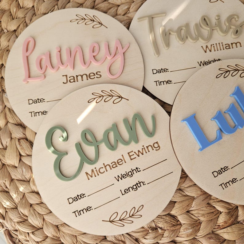May include: Four circular wooden birth announcement plaques featuring raised acrylic names in pink, sage green, and blue. Each plaque includes laser-engraved text for Date, Time, Weight, and Length, along with decorative leaf illustrations at the top and bottom.