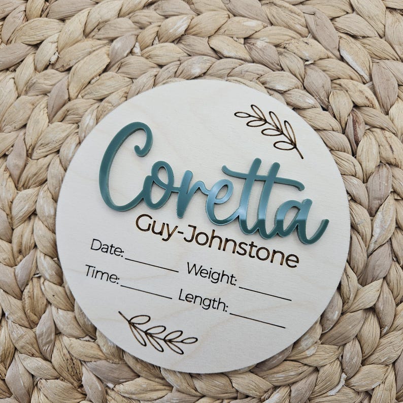 May include: A circular wooden birth announcement sign featuring the name Coretta Guy-Johnstone in raised teal acrylic lettering. The sign includes engraved lines for Date, Time, Weight, and Length, accented by two small engraved leaf sprig illustrations.