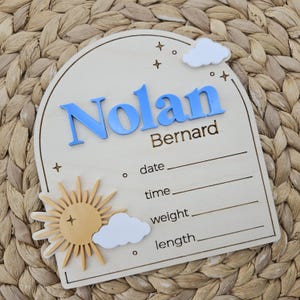 May include: An arched wooden birth announcement sign featuring the name Nolan in raised blue letters and Bernard in engraved text. The sign includes a yellow sun, white clouds, and engraved lines for date, time, weight, and length details.