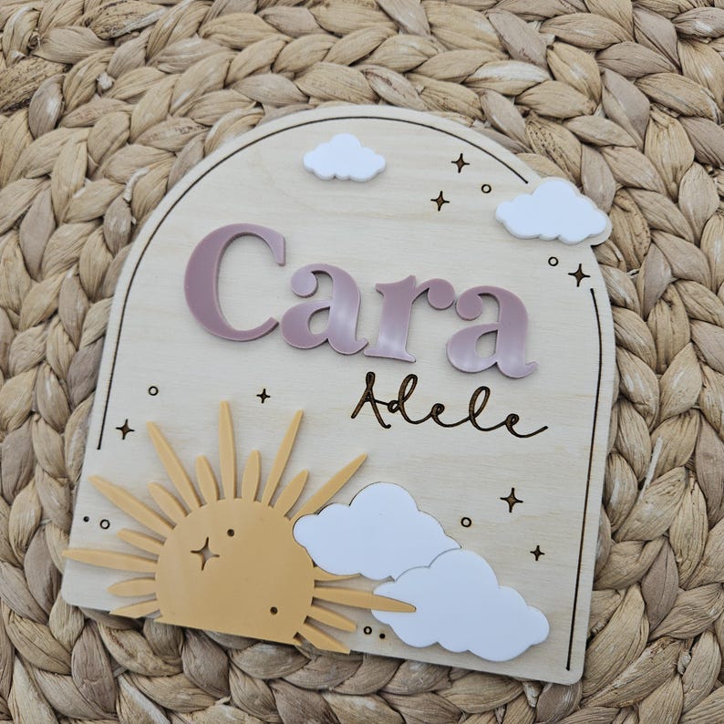 May include: An arched wooden plaque featuring the name Cara in raised mauve lettering and the name Adele in black script. The design includes a yellow sun, white clouds, and small star icons engraved on the light wood surface.