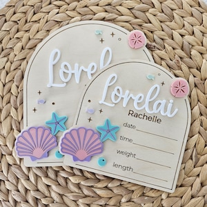 May include: Two wooden plaques with arched tops, featuring the name "Lorelei" in white script. One plaque includes the name "Rachelle" and space for date, time, weight, and length. Adorned with seashell, starfish, and sand dollar accents.