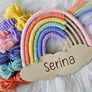 May include: A handmade rainbow macrame wall hanging with a wooden cloud featuring the name "Serina". The rainbow is crafted with pastel-colored yarn in shades of pink, orange, yellow, green, blue, and purple. Bundles of matching yarn are arranged around the rainbow.
