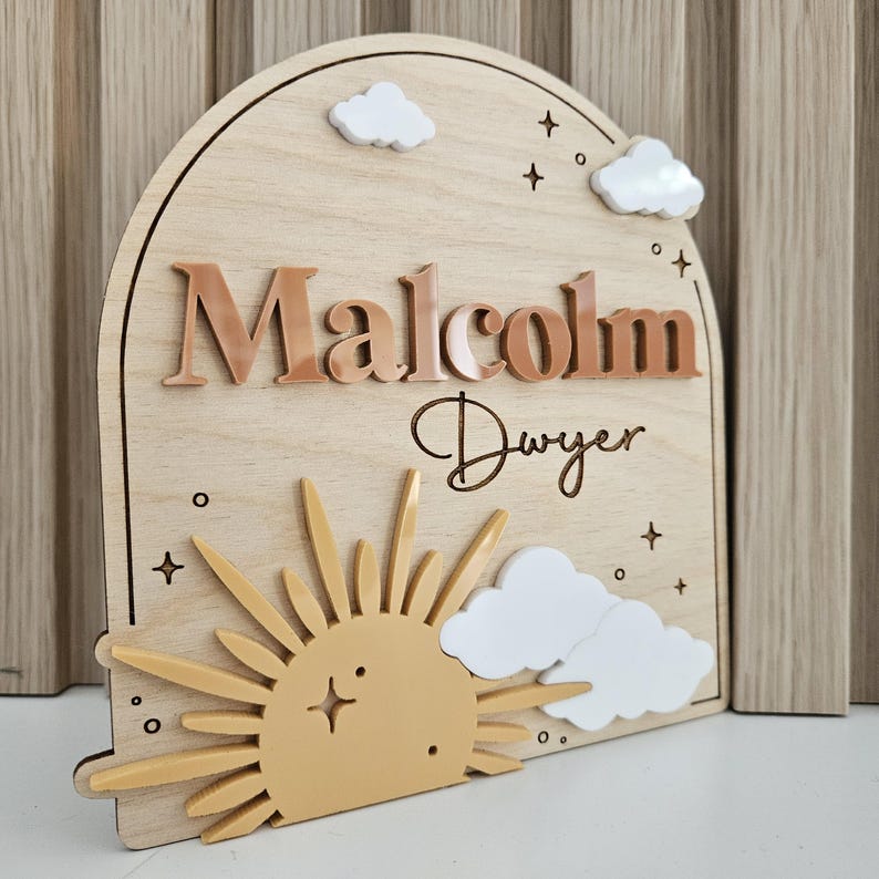 May include: An arched wooden sign featuring the name Malcolm Dwyer in raised brown lettering. The design includes a yellow sun with rays, white acrylic clouds, and small engraved stars and circles on a light wood grain background.