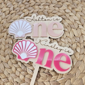 May include: Two wooden cake toppers featuring seashell designs and the text Ashton is one and Kinsley is one. The toppers have layered acrylic lettering in soft pink and bright pink, with white and pink seashell accents on a light wood base.