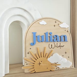 May include: An arched wooden sign featuring the name Julian in raised blue letters and the name Wilder in black script. The design includes a yellow sun, white clouds, and small star accents engraved on the light wood surface.