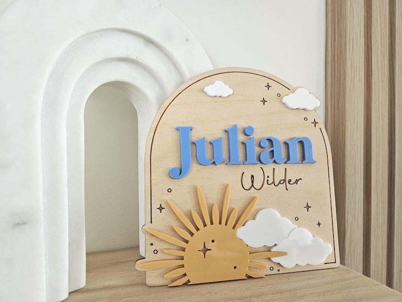 May include: An arched wooden sign featuring the name Julian in raised blue letters and the name Wilder in black script. The design includes a yellow sun, white clouds, and small star accents engraved on the light wood surface.