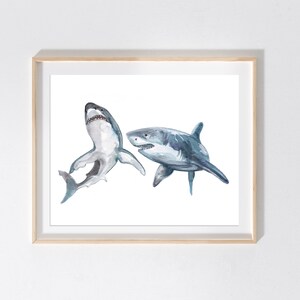 May include: Watercolor print of two sharks in shades of gray and blue. One shark is depicted with its mouth open, while the other is swimming to the right. The artwork is framed in a light wood frame, perfect for a nautical or ocean-themed decor.