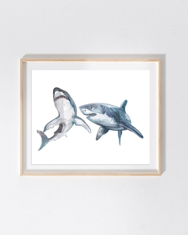 May include: Watercolor print of two sharks in shades of gray and blue. One shark is depicted with its mouth open, while the other is swimming to the right. The artwork is framed in a light wood frame, perfect for a nautical or ocean-themed decor.