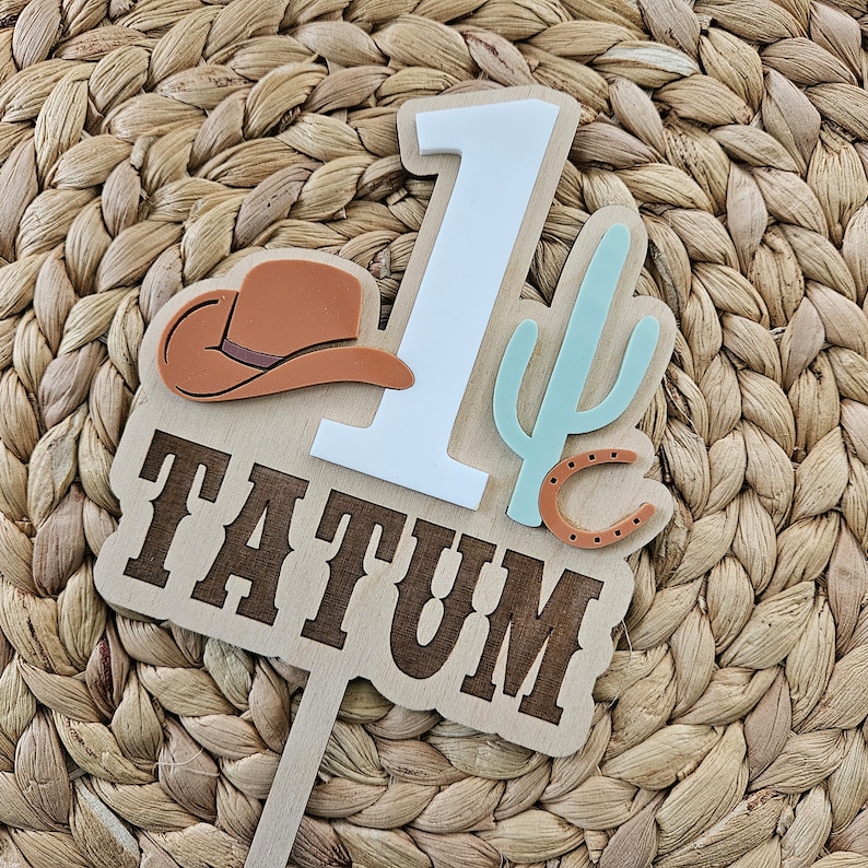 Peut inclure: D&eacute;coration de g&acirc;teau en bois sur le th&egrave;me du cow-boy. Elle pr&eacute;sente le nom "TATUM" en lettres marron, le chiffre blanc "1", un chapeau de cow-boy marron, un cactus vert clair et un fer &agrave; cheval. Con&ccedil;u pour un premier anniversaire.