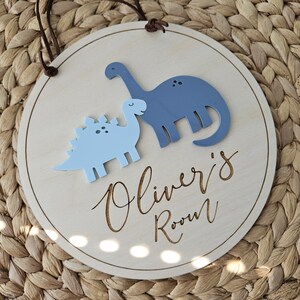 May include: A circular wooden sign featuring two blue dinosaur figures. The sign is inscribed with "Oliver's Room" in a cursive script. A brown leather cord is attached for hanging. The background has a woven texture.