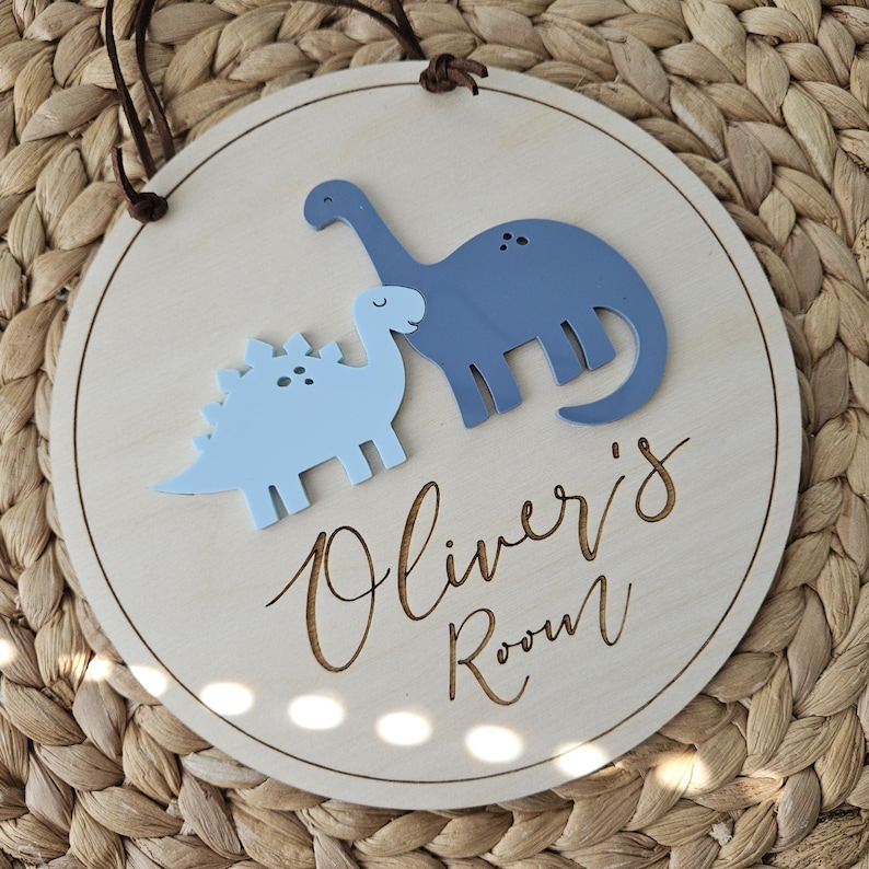 May include: A circular wooden sign featuring two blue dinosaur figures. The sign is inscribed with "Oliver's Room" in a cursive script. A brown leather cord is attached for hanging. The background has a woven texture.