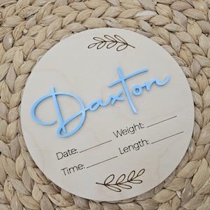May include: A circular light-colored wooden plaque featuring the name Daxton in raised light blue script lettering. The plaque includes engraved leaf motifs at the top and bottom, with blank lines for recording Weight, Date, Length, and Time.
