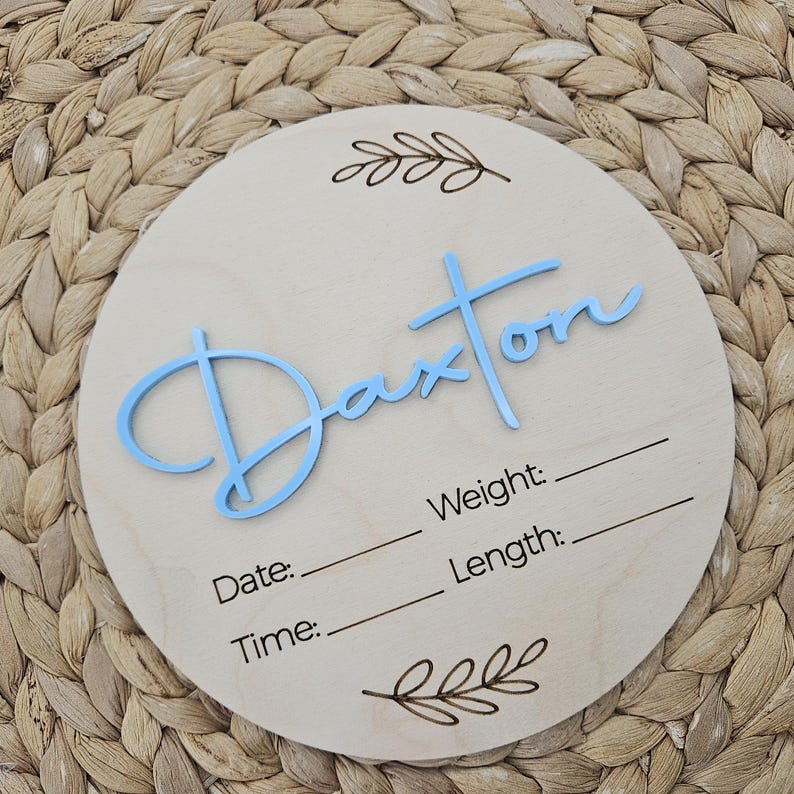 May include: A circular light-colored wooden plaque featuring the name Daxton in raised light blue script lettering. The plaque includes engraved leaf motifs at the top and bottom, with blank lines for recording Weight, Date, Length, and Time.
