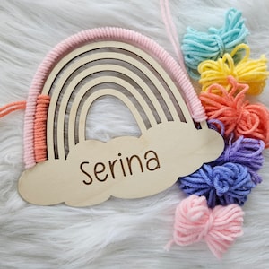 May include: Wooden rainbow-shaped wall decor with a cloud-shaped base. The rainbow features a macrame design in pink and peach, with a name engraved on the cloud. Colorful yarn pom-poms in shades of blue, yellow, orange, purple, and pink are attached.