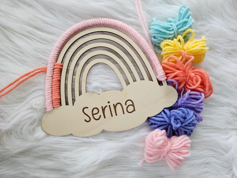 May include: Wooden rainbow-shaped wall decor with a cloud-shaped base. The rainbow features a macrame design in pink and peach, with a name engraved on the cloud. Colorful yarn pom-poms in shades of blue, yellow, orange, purple, and pink are attached.