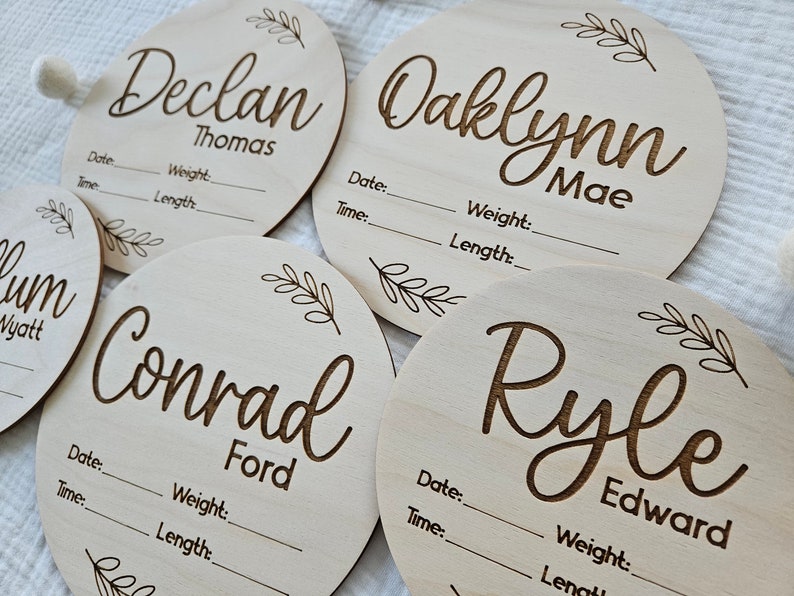 May include: Four circular light-colored wooden plaques engraved with names and birth details including Date, Time, Weight, and Length. Each plaque features a decorative leaf sprig design. The names Declan Thomas, Oaklynn Mae, Conrad Ford, and Ryle Edward are displayed in a script font.