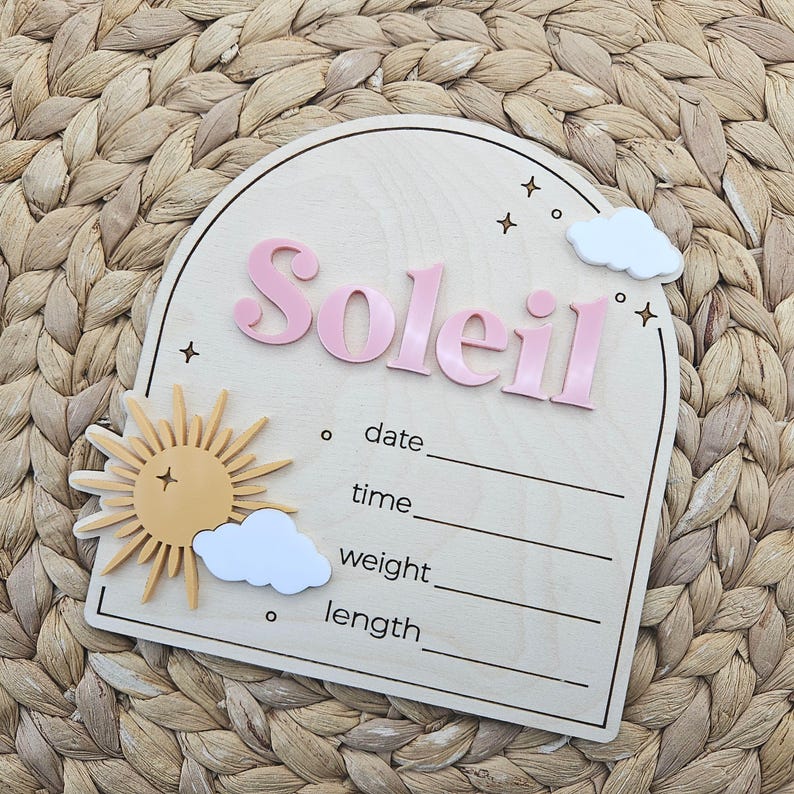 May include: An arched wooden birth announcement sign featuring the name Soleil in raised pink lettering. The sign includes a yellow sun icon, white clouds, and engraved stars. It has blank lines for date, time, weight, and length details.