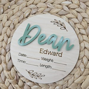 May include: A circular wooden birth announcement sign featuring the name Dean in raised sage green script, with the name Edward and the labels Date, Time, Weight, and Length engraved in black, accented by two small etched leaf branch designs.