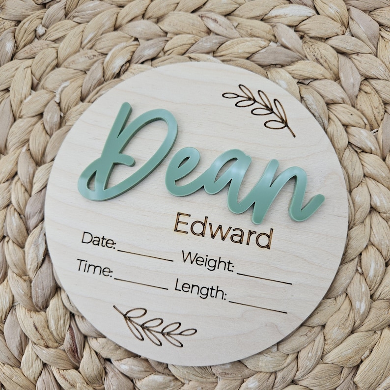 May include: A circular wooden birth announcement sign featuring the name Dean in raised sage green script, with the name Edward and the labels Date, Time, Weight, and Length engraved in black, accented by two small etched leaf branch designs.