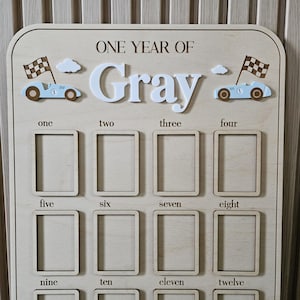 以下が含まれることがあります： 「ONE YEAR OF Gray」の文字が入った木製のマイルストーンボード。12個の写真スロット、レーシングカー、雲、1から12までの数字が特徴です。赤ちゃんの最初の年のための装飾品です。