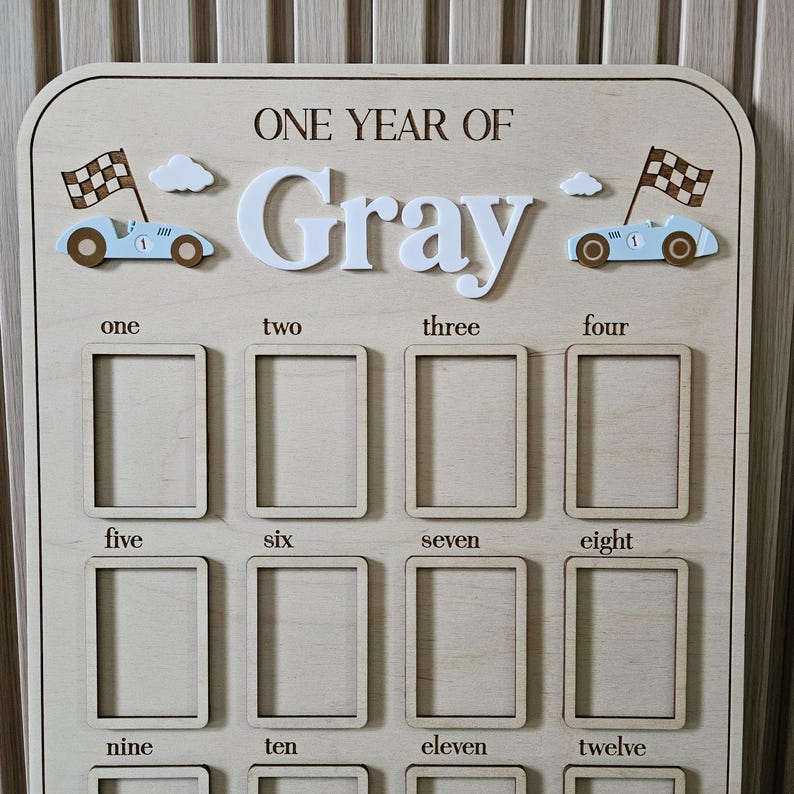 以下が含まれることがあります： 「ONE YEAR OF Gray」の文字が入った木製のマイルストーンボード。12個の写真スロット、レーシングカー、雲、1から12までの数字が特徴です。赤ちゃんの最初の年のための装飾品です。
