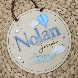 Op de afbeelding: Een rond houten wandbord met de naam Nolan in verhoogde blauwe letters en Joseph in cursief schrift. Het ontwerp bevat een lichtblauwe heteluchtballon, een lichtgroen vliegtuig, witte wolken en kleine zilveren sterren.