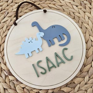 May include: A circular wooden sign with a light brown finish, featuring two dinosaur figures in light blue and blue. The name "ISAAC" is written in green letters below the dinosaurs. A brown cord is attached for hanging.