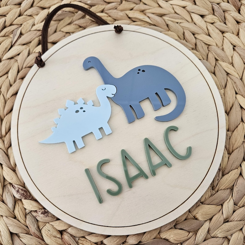 May include: A circular wooden sign with a light brown finish, featuring two dinosaur figures in light blue and blue. The name "ISAAC" is written in green letters below the dinosaurs. A brown cord is attached for hanging.