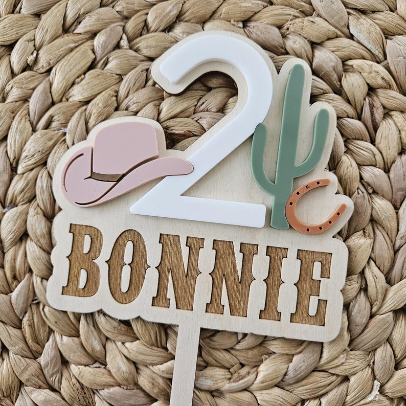 Peut inclure: Un cake topper en bois sur le th&egrave;me du cow-boy. Le topper pr&eacute;sente le chiffre 2 en blanc, un chapeau de cow-boy rose, un cactus vert et un fer &agrave; cheval marron. Le nom "BONNIE" est affich&eacute; en lettres de bois.
