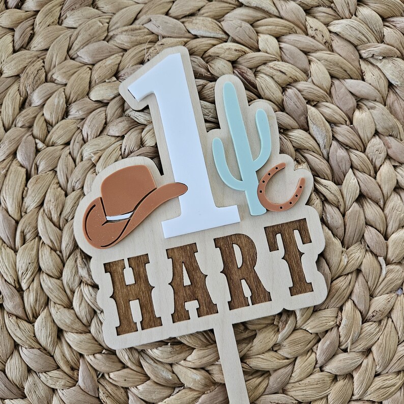Peut inclure: Un cake topper en bois sur le th&egrave;me du cow-boy. Le topper pr&eacute;sente le chiffre 1 en blanc, un chapeau de cow-boy marron, un cactus bleu clair et le nom "HART" en lettres marron. Un fer &agrave; cheval est &eacute;galement inclus.