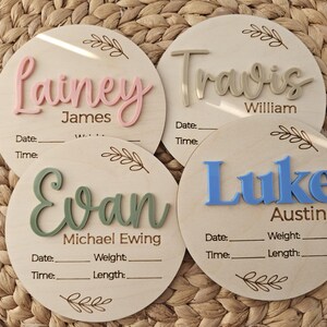May include: Four circular wooden birth announcement plaques featuring raised 3D lettering in pink, beige, green, and blue. Each plaque includes engraved text for names, date, time, weight, and length, accented with a delicate leaf branch design.