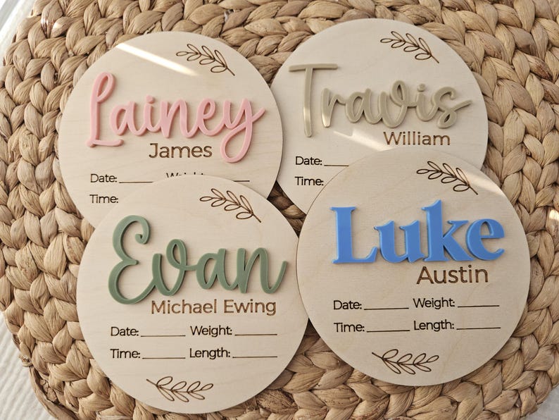 May include: Four circular wooden birth announcement plaques featuring raised 3D lettering in pink, beige, green, and blue. Each plaque includes engraved text for names, date, time, weight, and length, accented with a delicate leaf branch design.