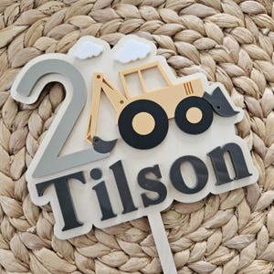 Könnte beinhalten: Ein personalisierter Tortenstecker mit einer großen grauen Zahl 2, einem beigen Bagger und zwei weißen Wolken. Der Name Tilson ist in fetten, dunkelgrauen Buchstaben unten angebracht. Das Design liegt auf einer hellen Holzbasis mit Stiel.