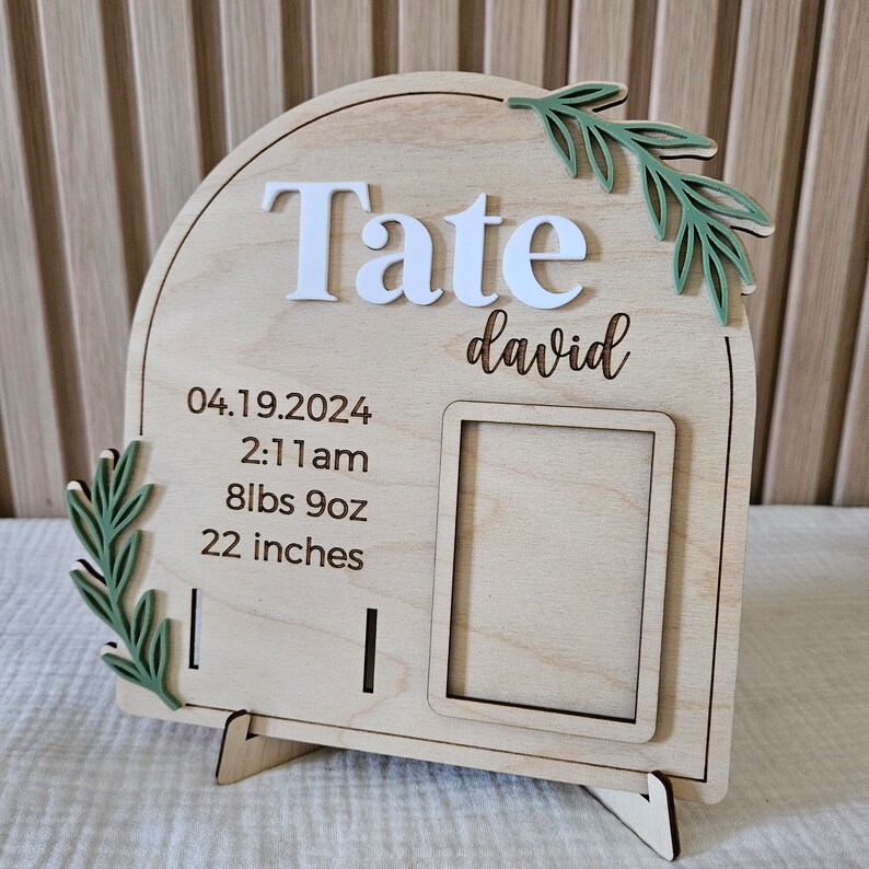 May include: A wooden birth announcement sign with the name "Tate" in white letters and "david" in script. The sign includes the birth date, time, weight (8lbs 9oz), and length (22 inches). Green leaf accents are on the top and left side.