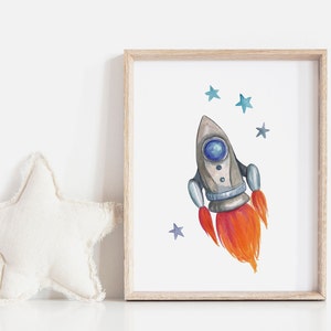 May include: Watercolor illustration of a gray and blue rocket ship with orange flames, framed in a light wood frame. Blue stars are scattered around the rocket on a white background. A white star-shaped pillow is on the left.