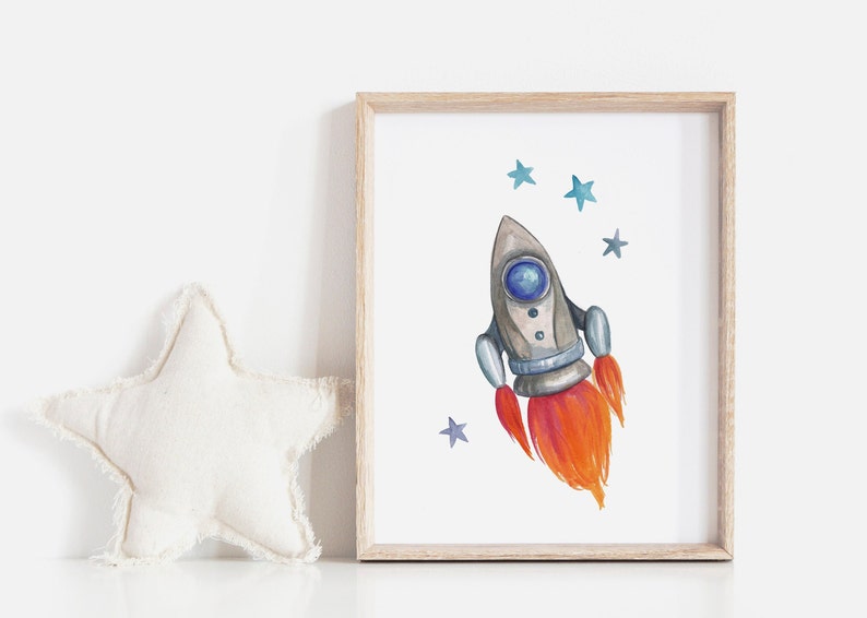 May include: Watercolor illustration of a gray and blue rocket ship with orange flames, framed in a light wood frame. Blue stars are scattered around the rocket on a white background. A white star-shaped pillow is on the left.