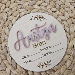 May include: A circular wooden birth announcement sign featuring the name Austyn in raised mauve script lettering, with the name Bren and the labels Date, Time, Weight, and Length laser-engraved below, accented by two decorative leaf illustrations.