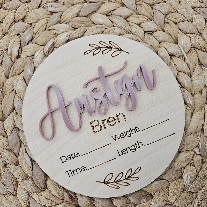 May include: A circular wooden birth announcement sign featuring the name Austyn in raised mauve script lettering, with the name Bren and the labels Date, Time, Weight, and Length laser-engraved below, accented by two decorative leaf illustrations.