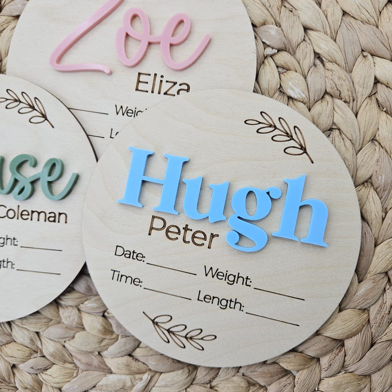 May include: Three circular wooden birth announcement plaques featuring raised acrylic names in pink, green, and blue. Each plaque includes laser-engraved text for Date, Time, Weight, and Length, along with decorative leaf illustrations at the top and bottom.