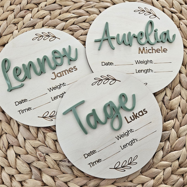 May include: Three circular wooden birth announcement signs with light wood grain. Each features raised sage green script names, brown printed middle names, and black engraved lines for Date, Time, Weight, and Length, accented by small leaf illustrations.