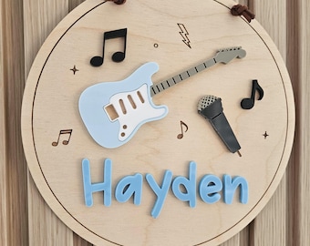 May include: A circular wooden wall plaque featuring a light blue electric guitar, a grey microphone, and black musical notes. The name Hayden is displayed in raised light blue letters at the bottom. The design includes engraved lightning bolts and stars.