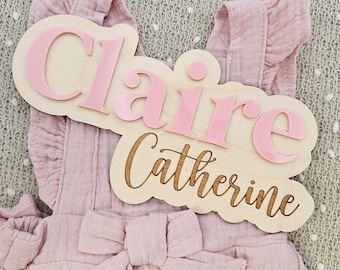 May include: A wooden name sign with the name "Claire" in pink acrylic letters, and the name "Catherine" in a script font. The sign is placed on a pink ruffled romper with a bow.