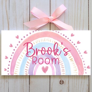 May include: A rectangular sign featuring a watercolour rainbow design in pink, blue, and gold. The sign says "Brook's Room" in pink script, with small pink hearts. A pink ribbon is attached at the top for hanging.