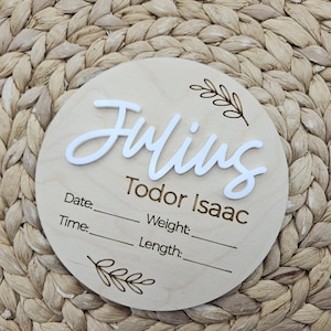 May include: A circular wooden birth announcement sign featuring the name Julius in raised white script lettering and the name Todor Isaac in engraved text. The sign includes engraved fields for Date, Time, Weight, and Length with leaf accents.